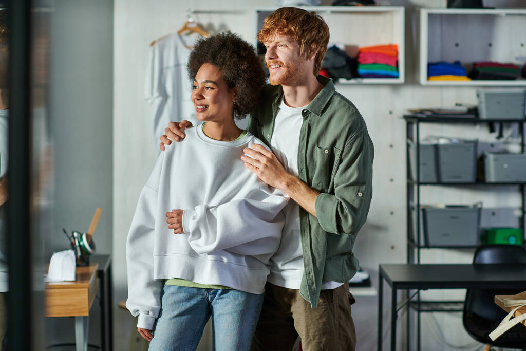 Χαρούμενη νεαρή κοκκινομάλλα τεχνίτη αγκαλιάζει αφροαμερικανή συνάδελφο και κοιτάζει μακριά ενώ στέκεται σε θολή στούντιο εκτύπωσης, συνεργατική ιδιοκτήτες επιχειρήσεων που εργάζονται μαζί  - Φωτογραφία, εικόνα