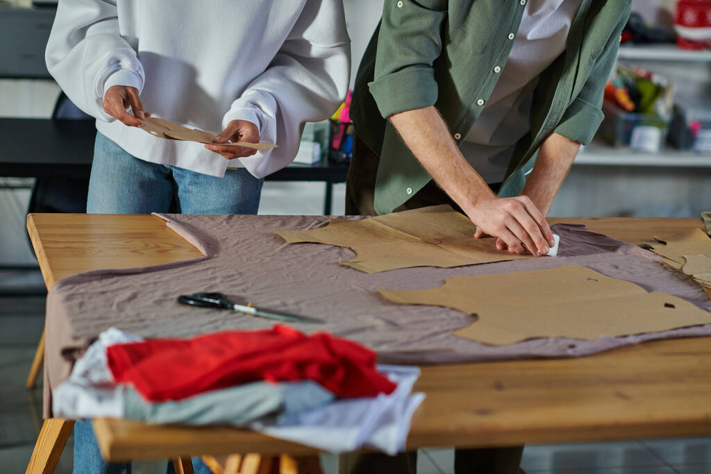 印刷スタジオでテーブルの上に布をマーキング同僚ながら、縫製パターンを保持しているアフリカ系アメリカ人の職人の作物ビュー、共同事業主が共同で作業  - 写真・画像