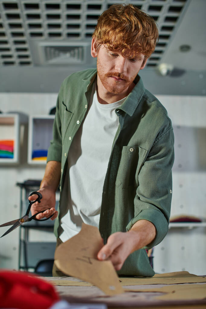 Young redhead craftsman holding scissors and blurred sewing pattern near fabric on table in blurred print studio at background, multitasking business owner managing multiple project - Photo, Image