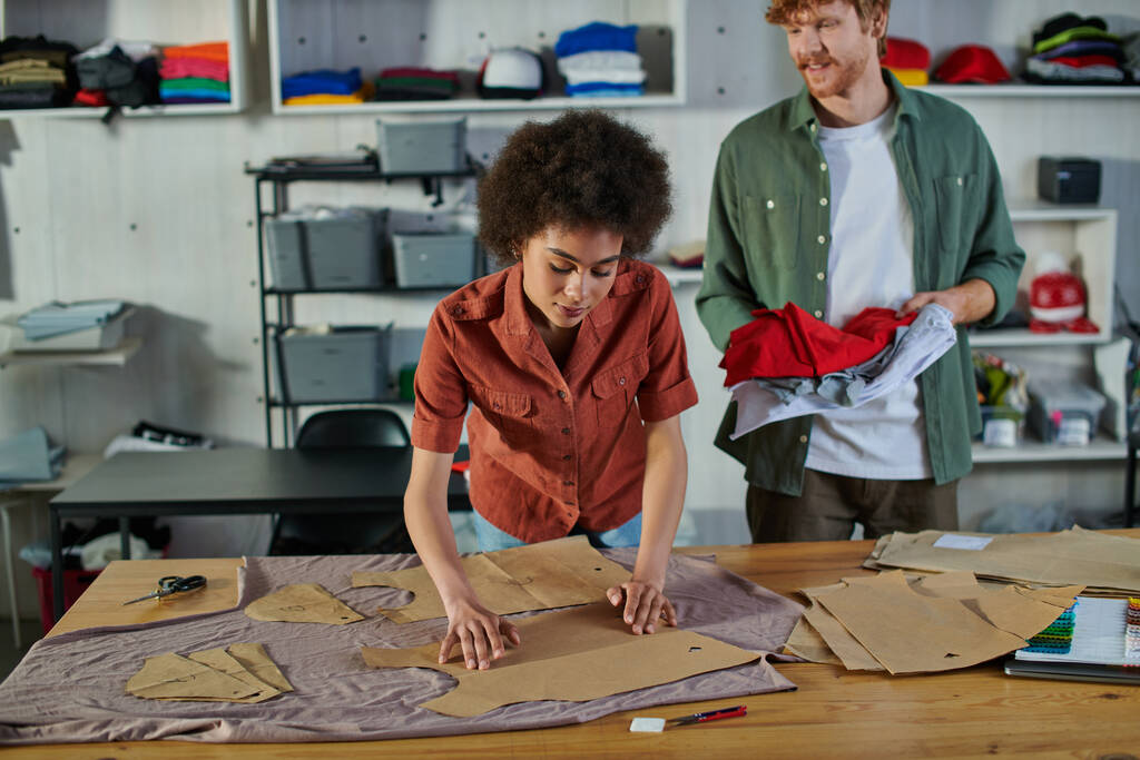 Jonge Afrikaans-Amerikaanse ambachtelijke vrouw zet naaipatronen op stof en werkt in de buurt van glimlachende roodharige collega in print studio, ambitieuze jonge ondernemers concept  - Foto, afbeelding