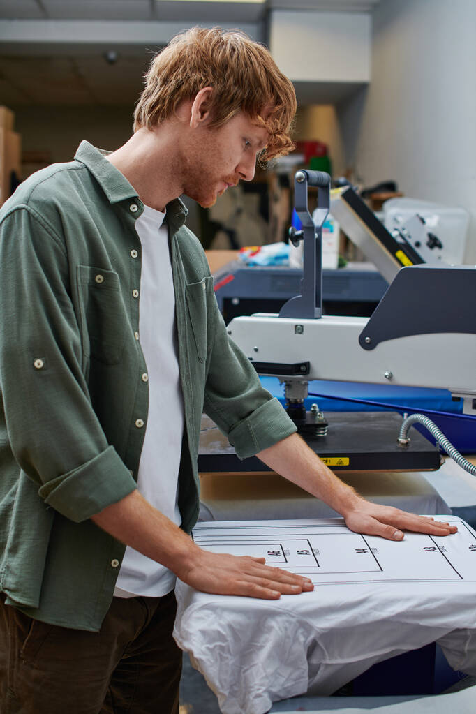 Вид збоку молодого рудого майстра, який працює з футболкою та екранним друкарським верстатом у розмитій друкарській студії на фоні, концепція малого бізнесу, орієнтована на клієнта - Фото, зображення