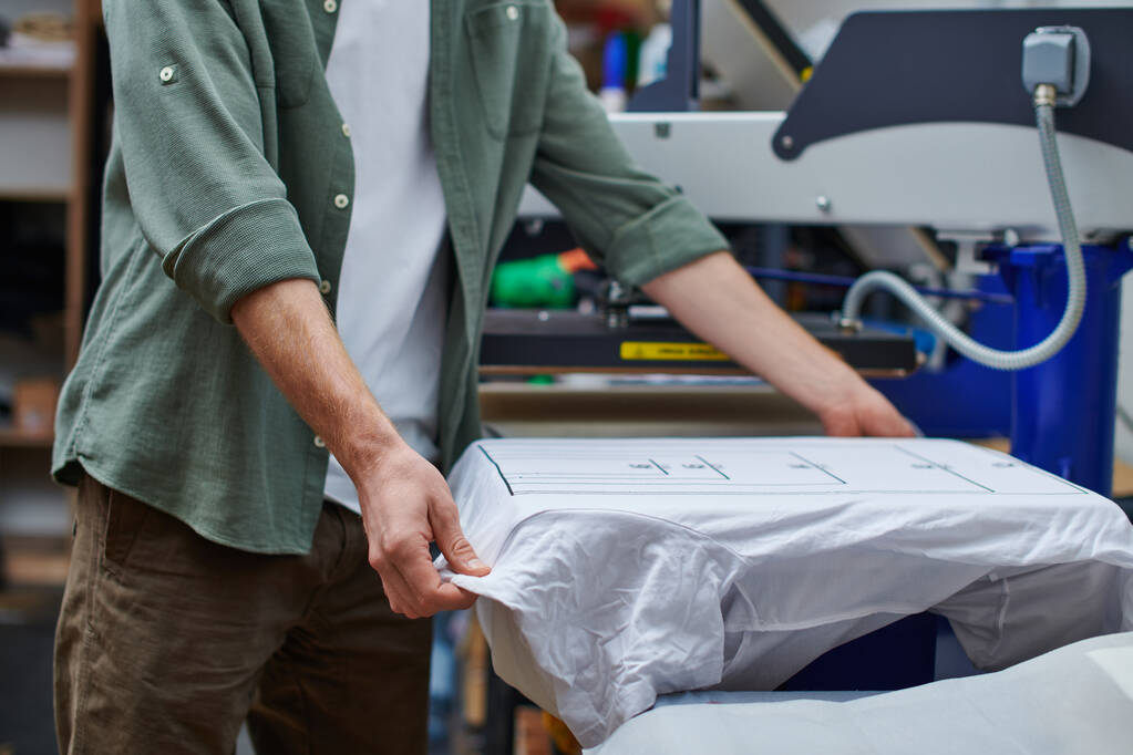 Widok na młodego rzemieślnika trzymającego koszulkę z oznakowaniem podczas pracy z maszyną do sitodruku w pracowni druku w tle, zorientowanej na klienta koncepcji małych firm - Zdjęcie, obraz