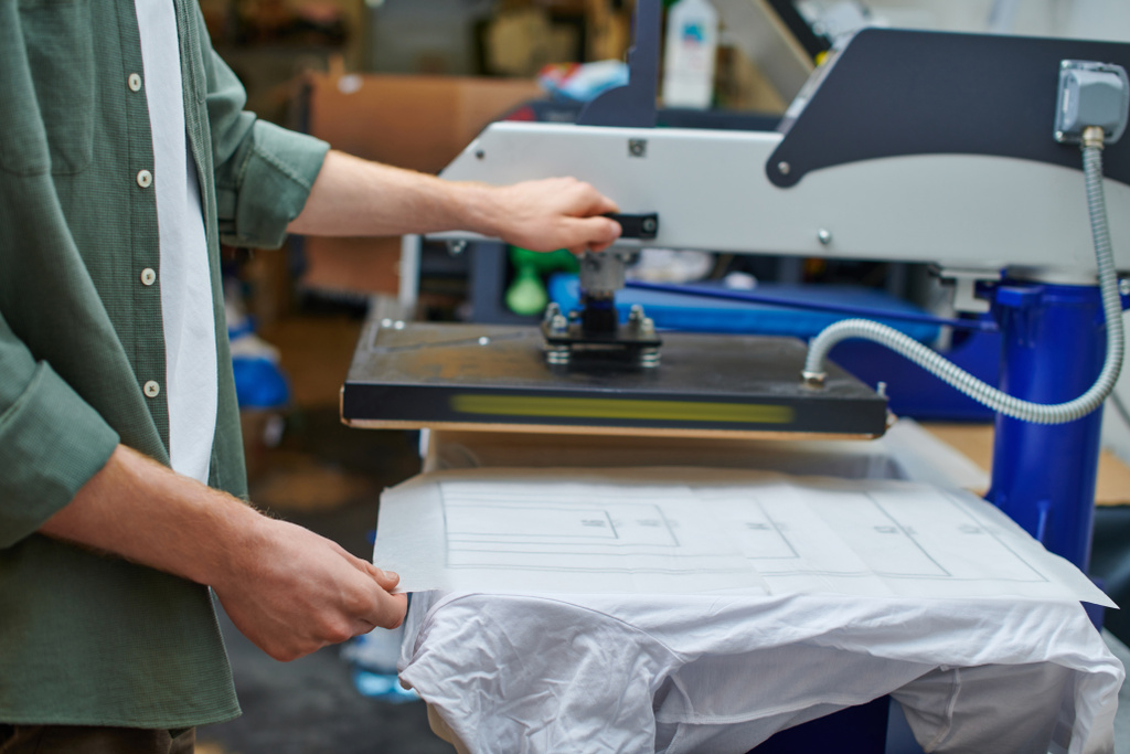 Oříznutý pohled na řemeslníka držícího vrstvu na tričku a pracující se sítotiskovým strojem v rozmazané dílně, zákaznicky zaměřený koncept malého podnikání - Fotografie, Obrázek