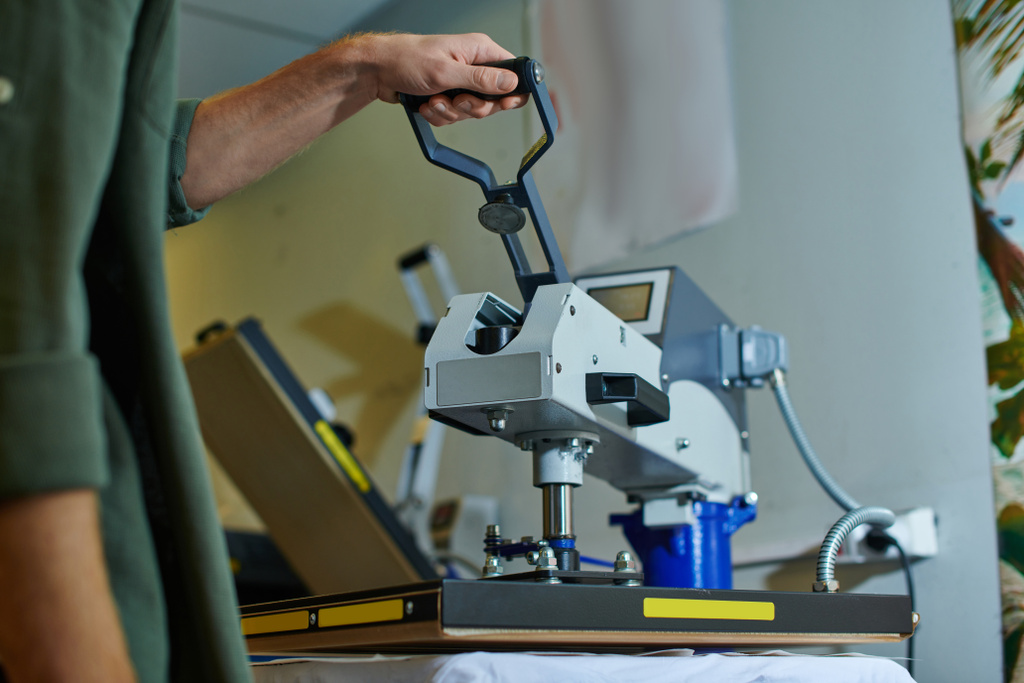 Καλλιεργημένη άποψη του νεαρού τεχνίτη που χρησιμοποιεί μηχάνημα εκτύπωσης οθόνης σε ρούχα ενώ εργάζεται σε θολό στούντιο εκτύπωσης στο παρασκήνιο, με επίκεντρο τον πελάτη έννοια της μικρής επιχείρησης - Φωτογραφία, εικόνα