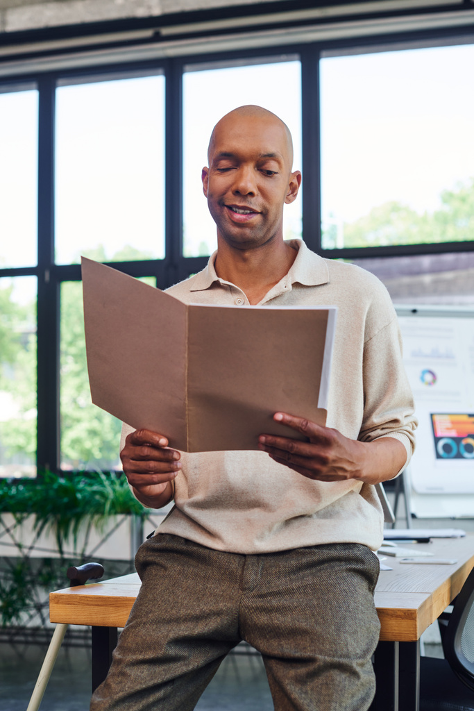 happy dark skinned man with myasthenia gravis disease holding folder and standing near desk and walking cane, bold african american office worker with ptosis eye syndrome, inclusion, work  - Photo, Image
