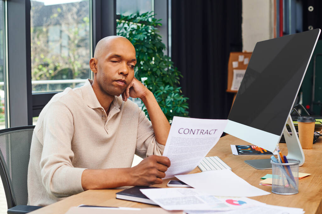 Kapsama, miastenya hastalığı olan Afro-Amerikan bir adam sözleşmeye bakıyor, koyu tenli ofis çalışanı masasında oturuyor, monitör, akıllı telefon, belgeler ve grafikler, ptosis sendromu.  - Fotoğraf, Görsel