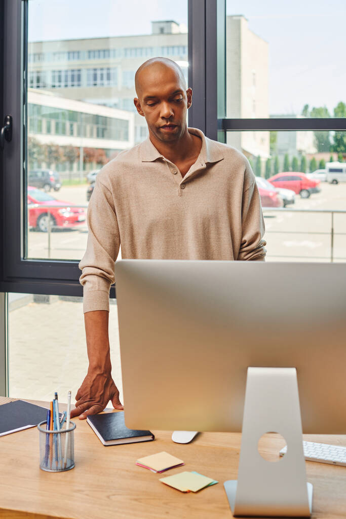 синдром міастенії гравіс, сміливий афроамериканський бізнесмен з синдромом Птозіса, який дивиться на комп'ютерний монітор, темношкірий офісний працівник в повсякденному вбранні, стоячи за столом, включення  - Фото, зображення