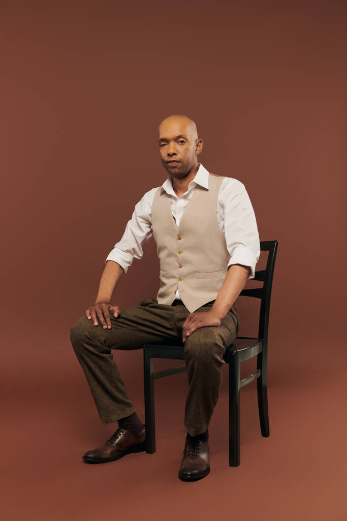 inclusión, hombre negrita de piel oscura con síndrome de miastenia gravis, sentado en la silla y mirando a la cámara, hombre afroamericano con enfermedad crónica sobre fondo marrón - Foto, imagen