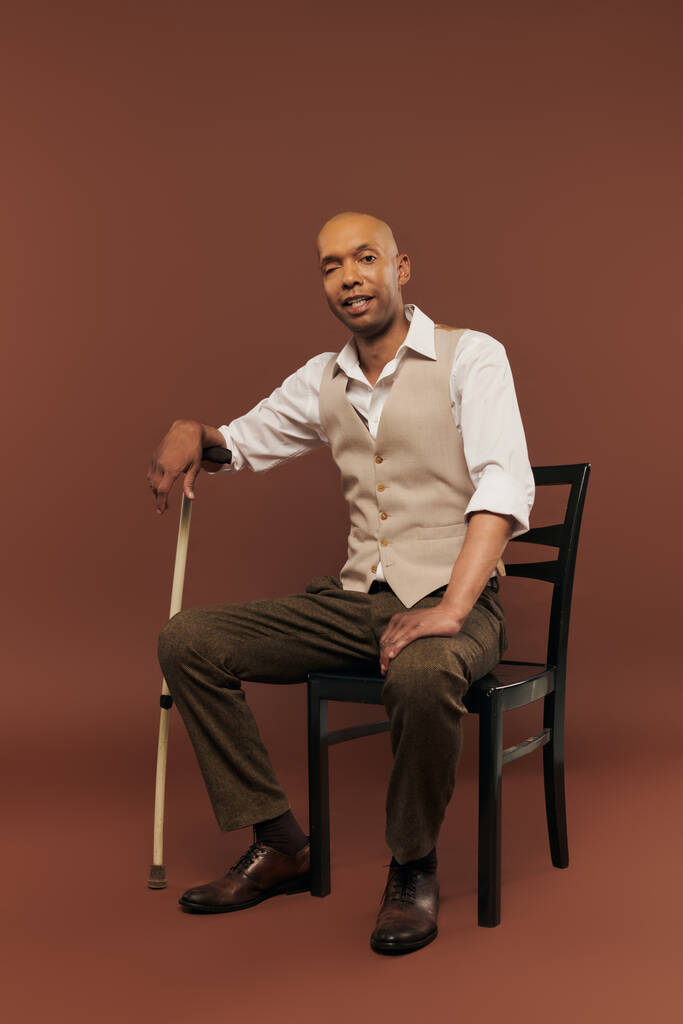 inclusion, real people, african american man with myasthenia gravis syndrome sitting on chair and looking at camera, dark skinned man with chronic disease holding walking stick on brown background - Photo, Image