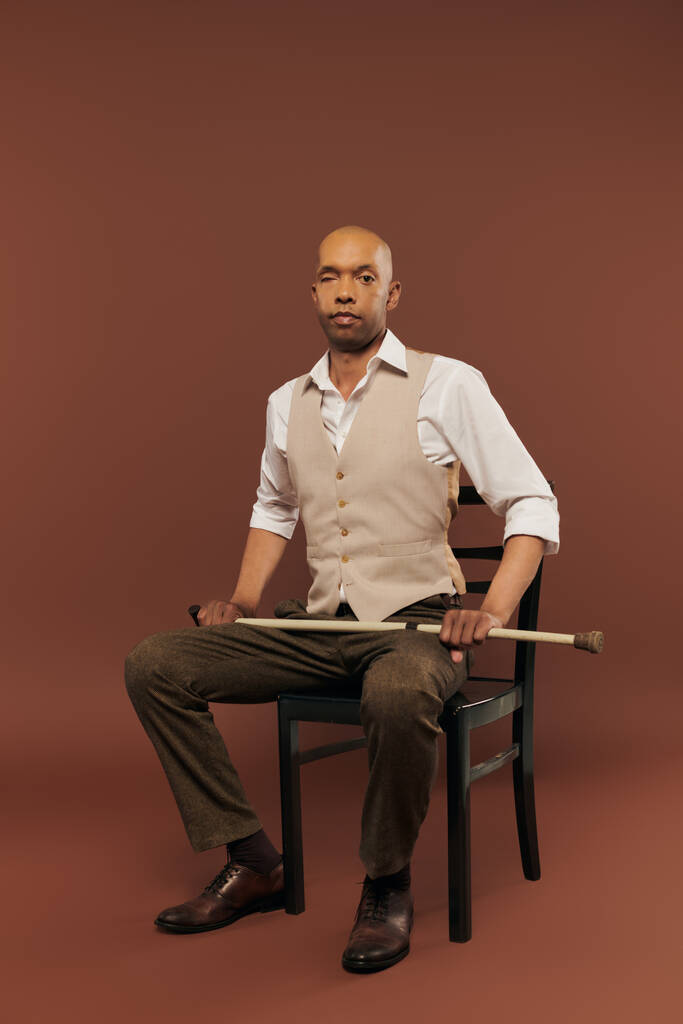 inclusion, homme afro-américain audacieux avec le syndrome de myasthénie grave, assis sur la chaise et regardant la caméra, homme à la peau foncée tenant la canne à pied sur fond brun, de vraies personnes  - Photo, image
