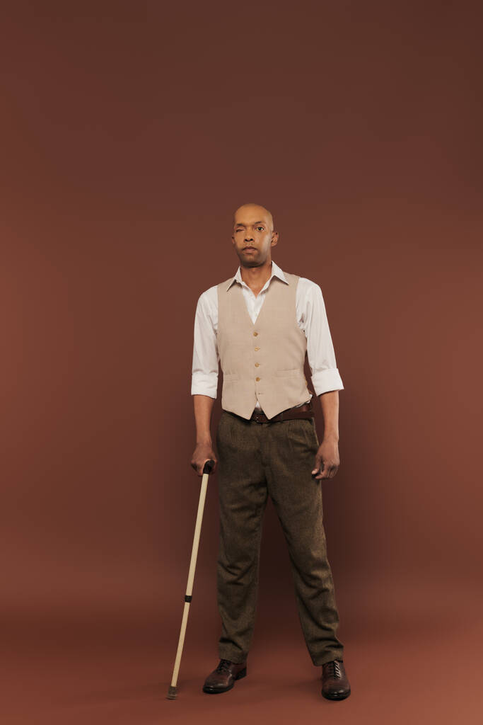 reale Menschen, mutiger afrikanisch-amerikanischer Mann mit Myasthenia-Gravis-Syndrom, stehend mit Gehstock auf braunem Hintergrund, stehend und in die Kamera blickend, Vielfalt und Inklusion, körperliche Beeinträchtigung  - Foto, Bild