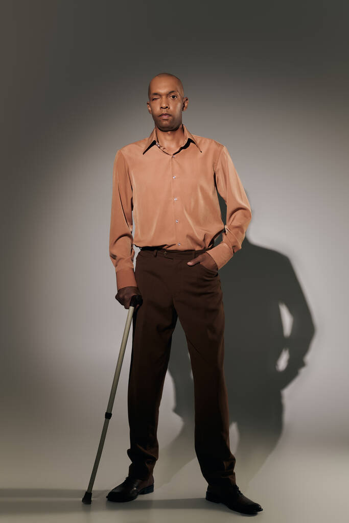 реальні люди, сміливий афроамериканський чоловік з міастенією, що стоїть з ходячою тростиною на сірому фоні, рука в кишені, поза, темношкіра людина в сорочці, різноманітність і включення, повна довжина  - Фото, зображення