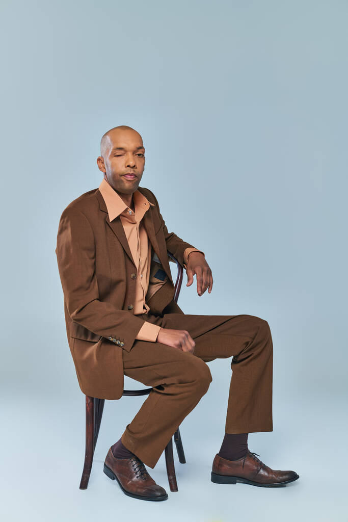 Invalidität, volle Länge des kräftigen afrikanisch-amerikanischen Mannes mit Myasthenia gravis, der auf einem Holzstuhl auf grauem Hintergrund sitzt, dunkelhäutige Person im Anzug, die in die Kamera schaut, Vielfalt und Inklusion  - Foto, Bild