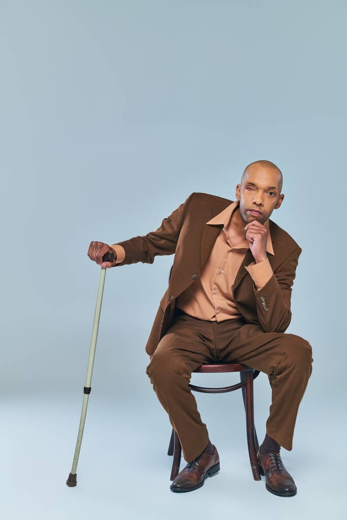 інвалідність, повнометражний сміливий афроамериканський чоловік з міастенією гравієм, що сидить на дерев'яному стільці на сірому фоні, темношкіра людина в костюмі, спираючись на прогулянкову тростину, різноманітність та включення  - Фото, зображення