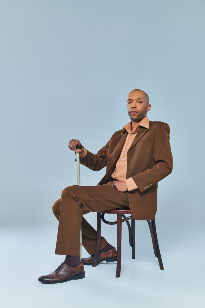 σύνδρομο πτώσης, πλήρες μήκος του τολμηρού Αφροαμερικανού άνδρα με μυασθένεια gravis κάθεται στην καρέκλα σε γκρι φόντο, σκούρο δέρμα άτομο με κοστούμι ακουμπά στο περπάτημα ζαχαροκάλαμο, την ποικιλομορφία και την ένταξη  - Φωτογραφία, εικόνα