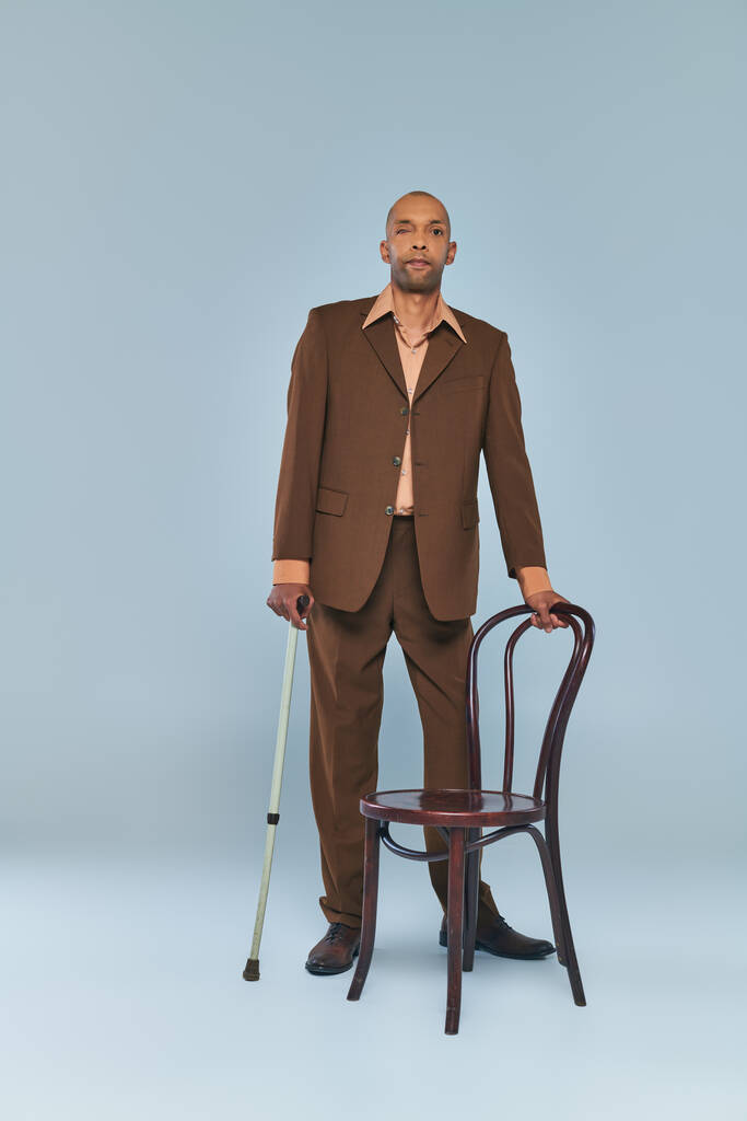 синдром птозу, повна довжина сміливого афроамериканця з міастенією гравієм, що стоїть біля стільця на сірому фоні, темношкіра людина в костюмі з ходячою тростиною, різноманітністю та включенням  - Фото, зображення