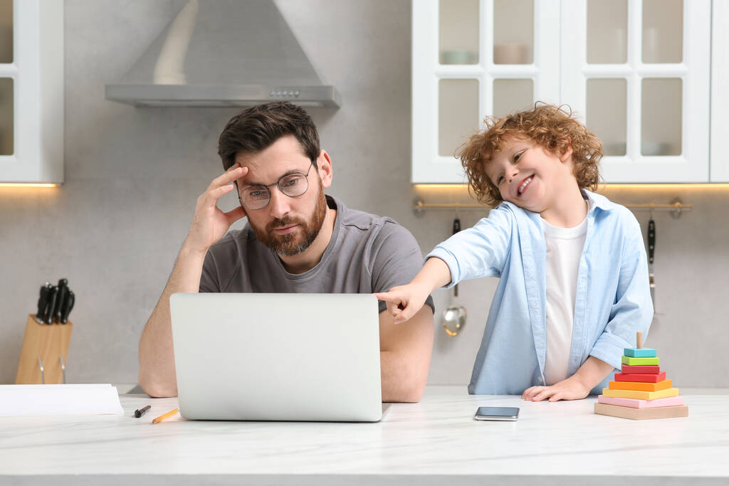 Pieni poika häiritsee isää, kun hän tekee etätyötä kotona. Mies kannettava tietokone ja hänen lapsensa pöydällä keittiössä - Valokuva, kuva