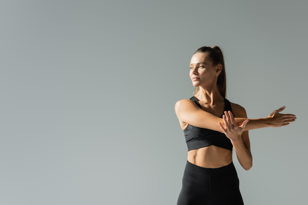 zelfverzekerde sportvrouw in sportbeha stretching armen en training geïsoleerd op grijze, actieve levensstijl  - Foto, afbeelding