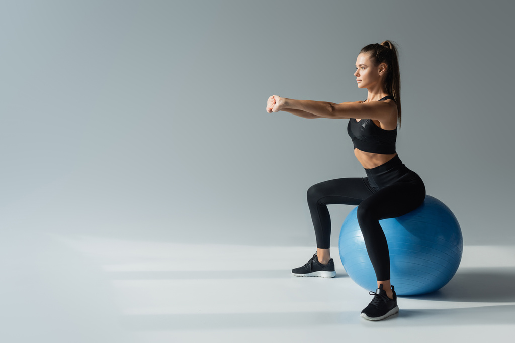 Siyah etekli esmer kadın gri arka planda fitness topu üzerinde egzersiz yapıyor, dengesini koruyor. - Fotoğraf, Görsel