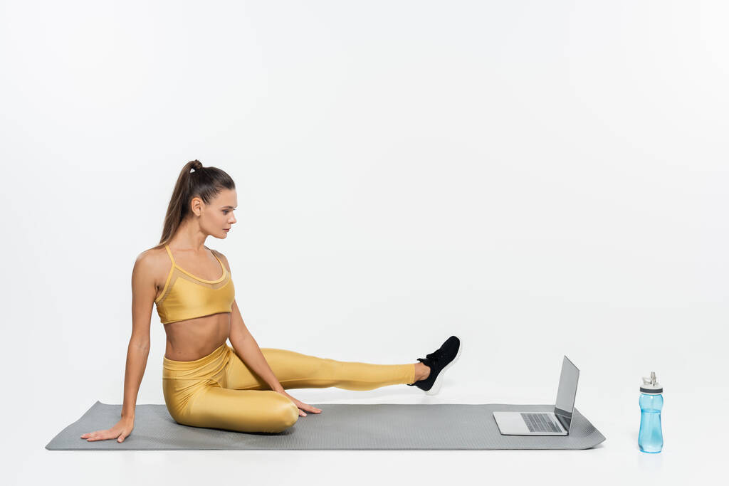 e-sports, mujer en ropa deportiva sentada cerca de la computadora portátil y la botella en la estera de fitness, fondo blanco - Foto, Imagen