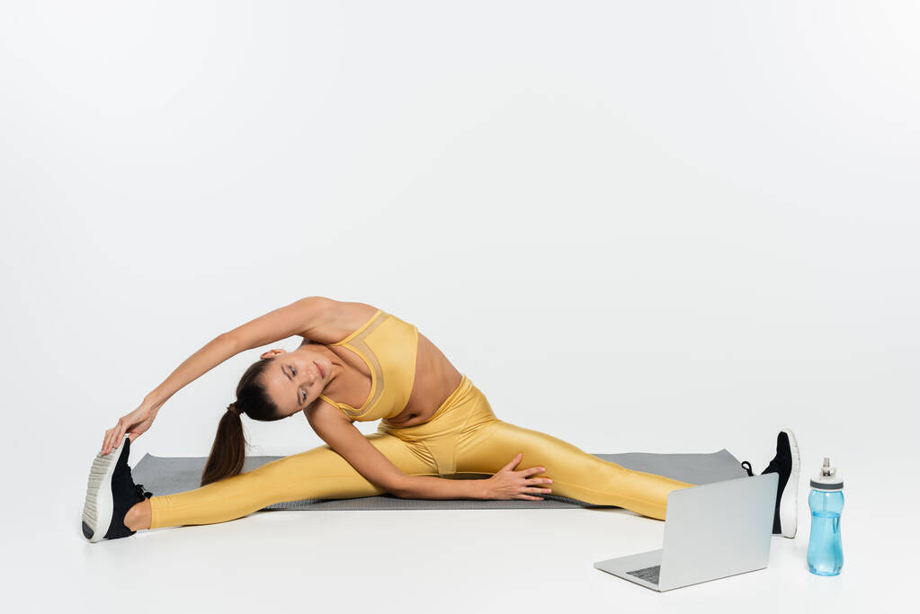 e-sport, vrouw in sportkleding stretching en kijken naar laptop op fitness mat op witte achtergrond  - Foto, afbeelding