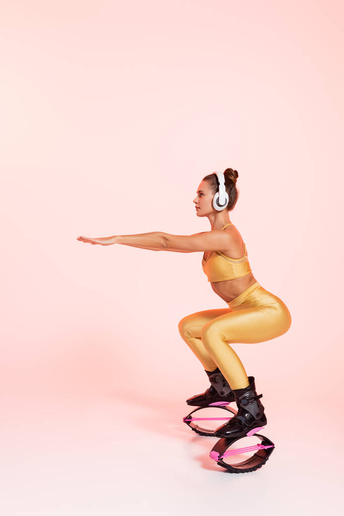 Denge, kangoo zıplayan ayakkabılı kadın ve pembe arka planda egzersiz yapan kablosuz kulaklıklar, çömelmeler. - Fotoğraf, Görsel