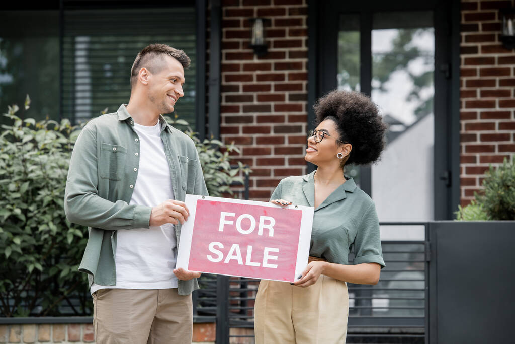 szczęśliwy wielonarodowych pośredników nieruchomości gospodarstwa na sprzedaż signboard, patrząc na siebie w pobliżu domu - Zdjęcie, obraz