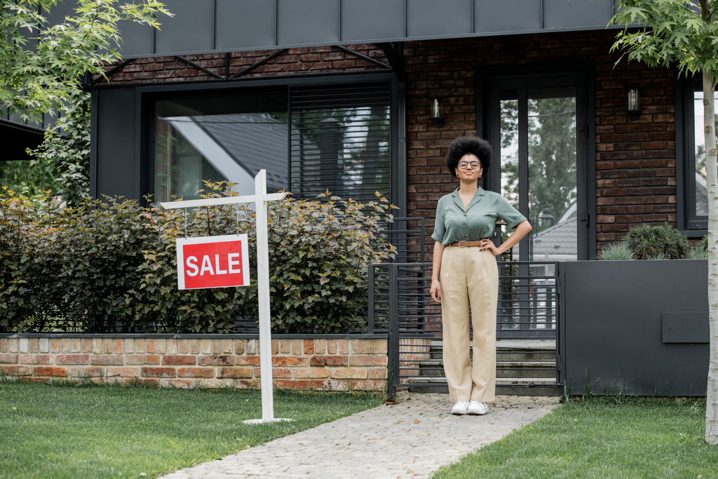 glücklich afrikanisch-amerikanischer Makler mit Hand auf Hüfte nahe Gebäude zum Verkauf in der Stadt - Foto, Bild