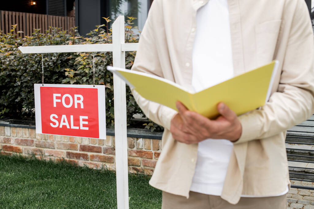 vue recadrée de l'agent immobilier debout avec dossier près de panneau à vendre en face de la maison de la ville - Photo, image