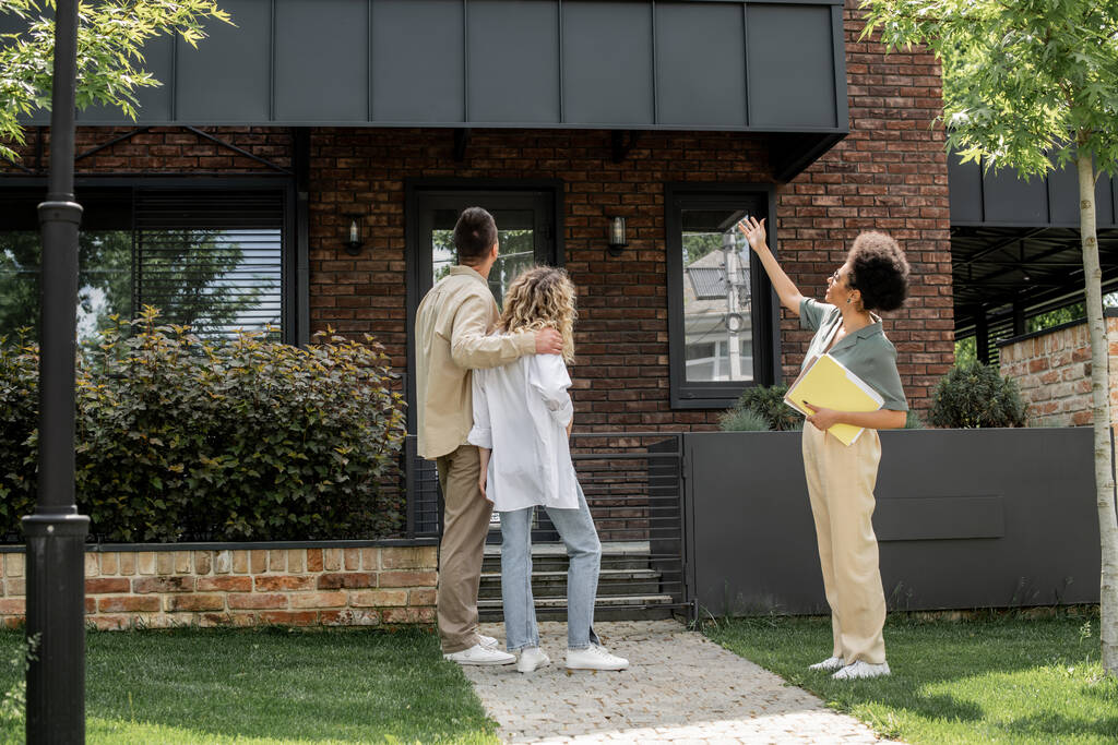 afroamericano agente de la propiedad señalando con la mano cerca de pareja abrazando junto a casa moderna - Foto, imagen