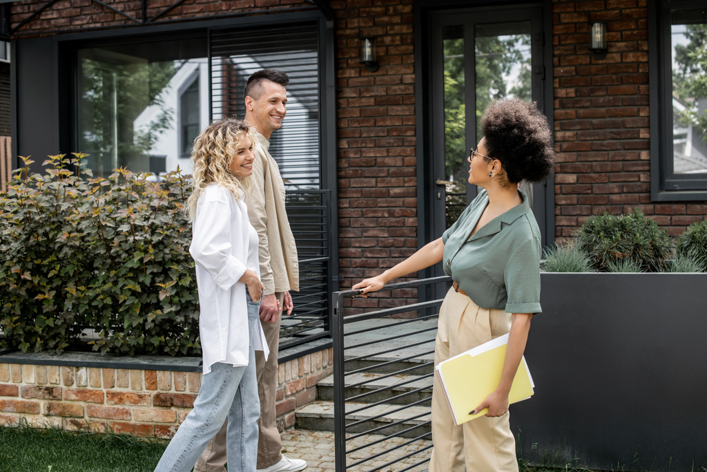 casal alegre conversando com corretor de imóveis afro-americano perto de edifício moderno na rua urbana - Foto, Imagem