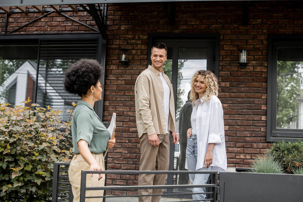 Afrikanisch-amerikanischer Immobilienmakler im Gespräch mit lächelndem Paar in der Nähe eines neuen modernen Häuschens zum Verkauf - Foto, Bild