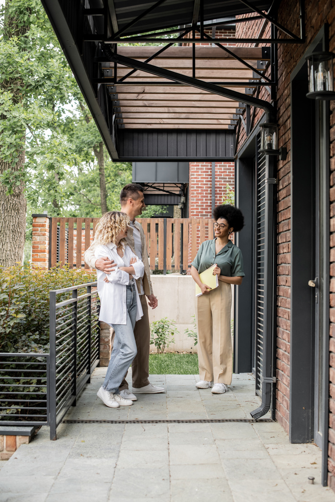 glimlachend Afrikaans Amerikaans makelaar met map praten met een paar nieuwe eigenaren in de buurt van huis - Foto, afbeelding
