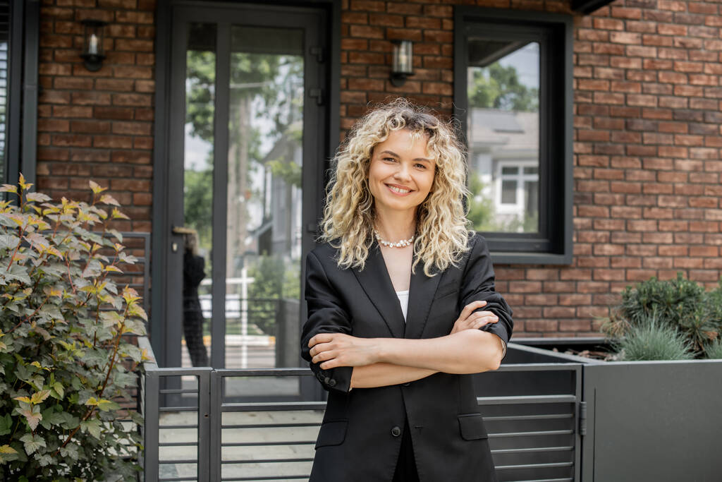 blonde and happy real estate agent standing with folded arms and looking at camera near cottage - Photo, Image