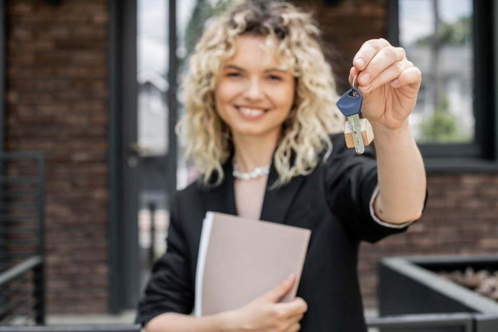 Размытый агент по недвижимости улыбается и смотрит в камеру, держа в руках папку и ключ от нового дома - Фото, изображение