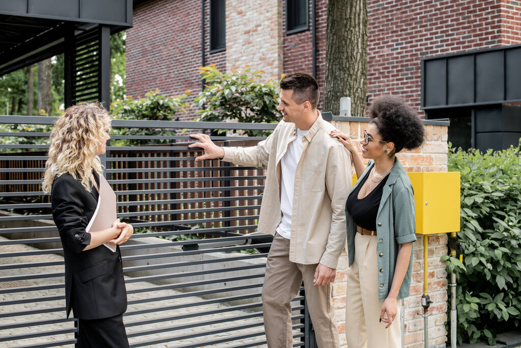 задоволена багатоетнічна пара, що стоїть біля агента з нерухомості і паркану нового міського котеджу - Фото, зображення