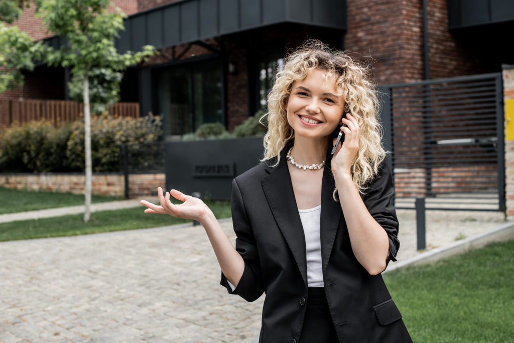 blonde property realtor pointing with hand and smiling at camera while talking on smartphone - Photo, Image
