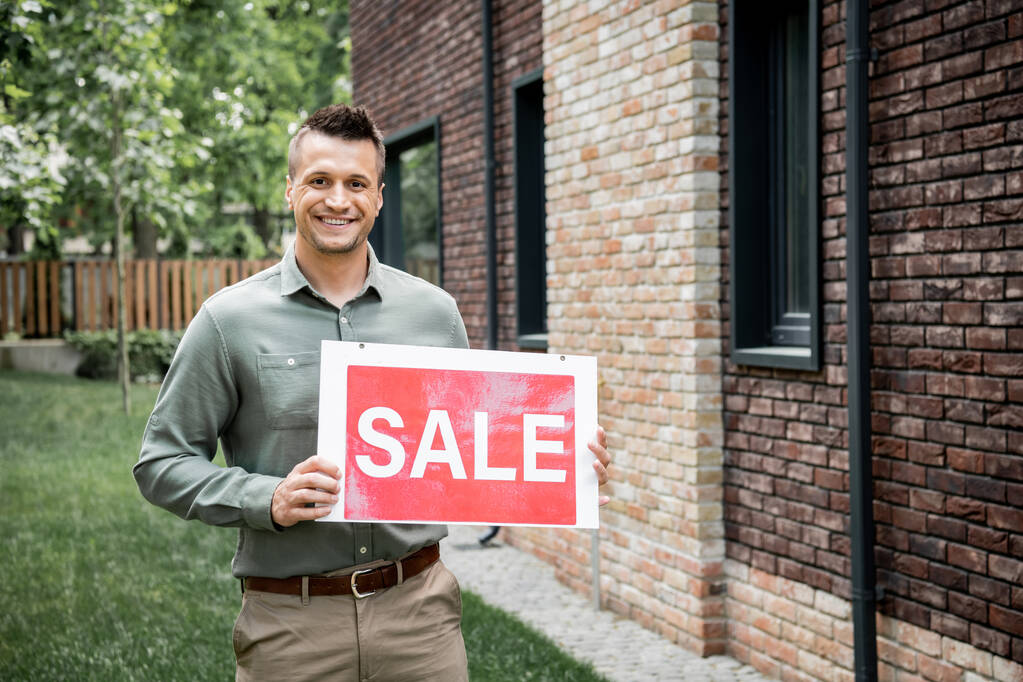 glücklich und zuversichtlich Immobilienmakler Blick auf Kamera und halten Verkauf Schild in der Nähe des neuen Hauses - Foto, Bild