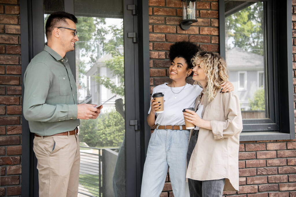 pośrednik nieruchomości rozmawia ze szczęśliwą międzyrasową parą lesbijek z papierowymi kubkami w pobliżu nowego domku - Zdjęcie, obraz