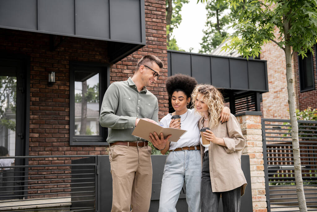 positive realtor showing documents to interracial lesbian couple with paper cups near house - Photo, Image