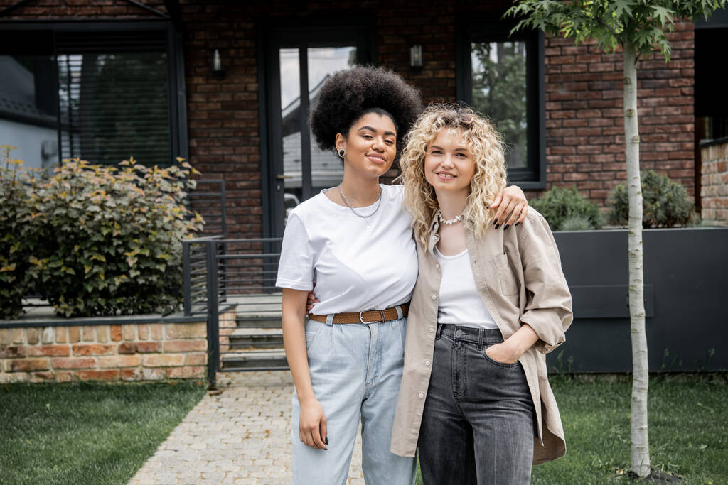 pleased multiethnic lesbian couple embracing and looking at camera near new private house - Photo, Image