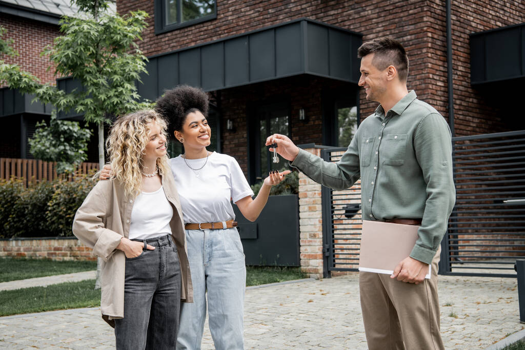 successful realtor with folder giving key from new house to pleased multiethnic lesbian couple - Photo, Image