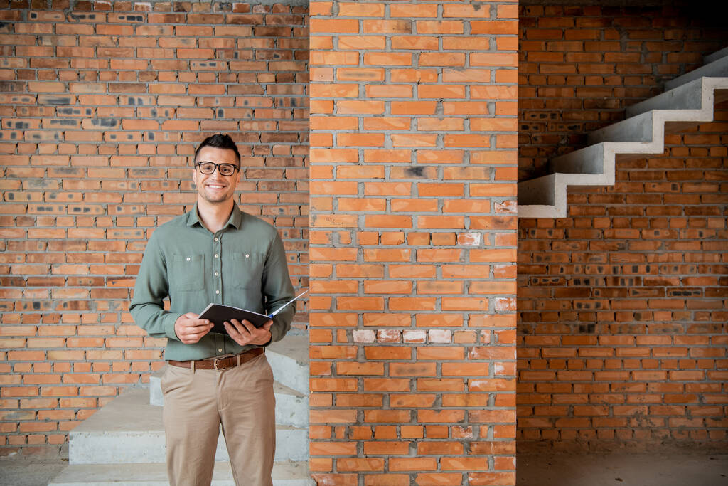 joyful real estate agent in eyeglasses looking at camera in house with unfinished interior - Photo, Image