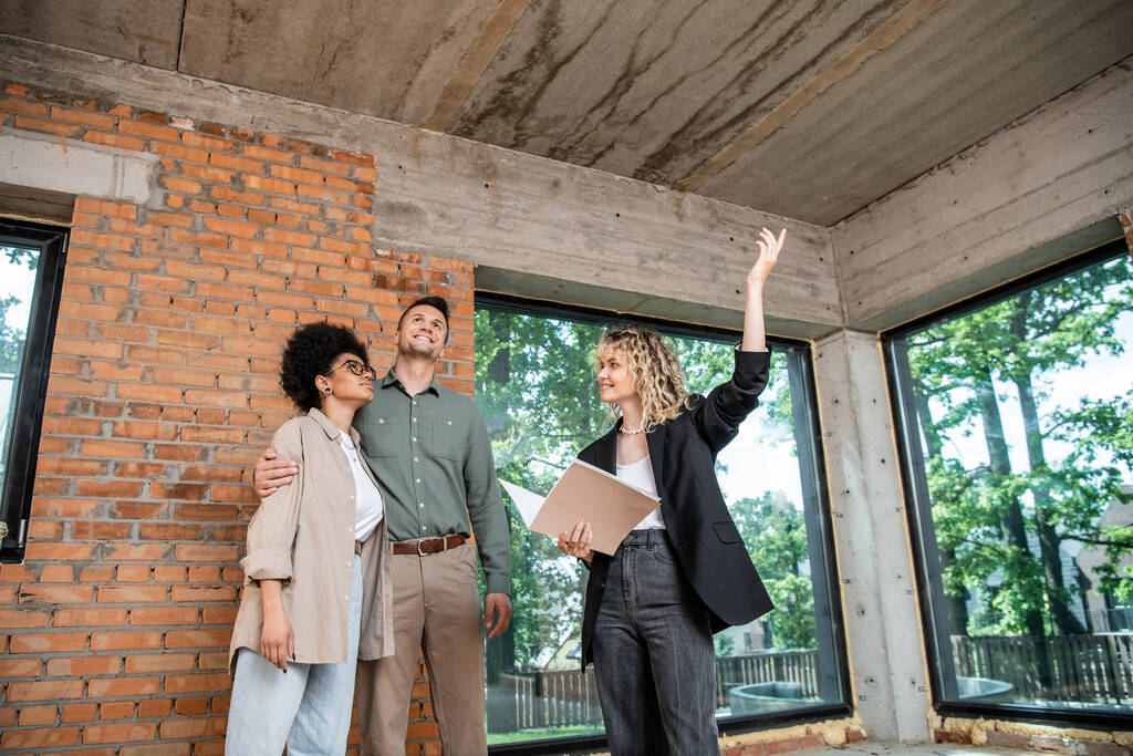 agente de bienes raíces apuntando hacia arriba con la mano en el techo mientras que muestra nueva casa a la pareja multiétnica - Foto, Imagen