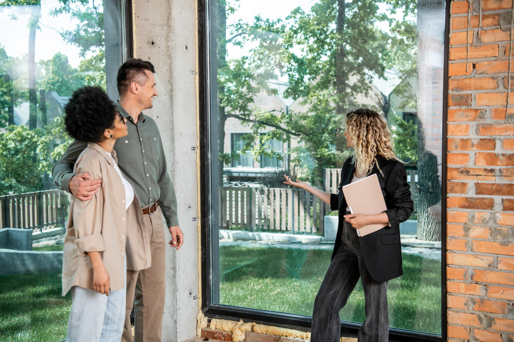 Immobilienmakler mit Ordner zeigt mit der Hand auf Fenster in neuem Ferienhaus in der Nähe gemischtrassiges Paar - Foto, Bild