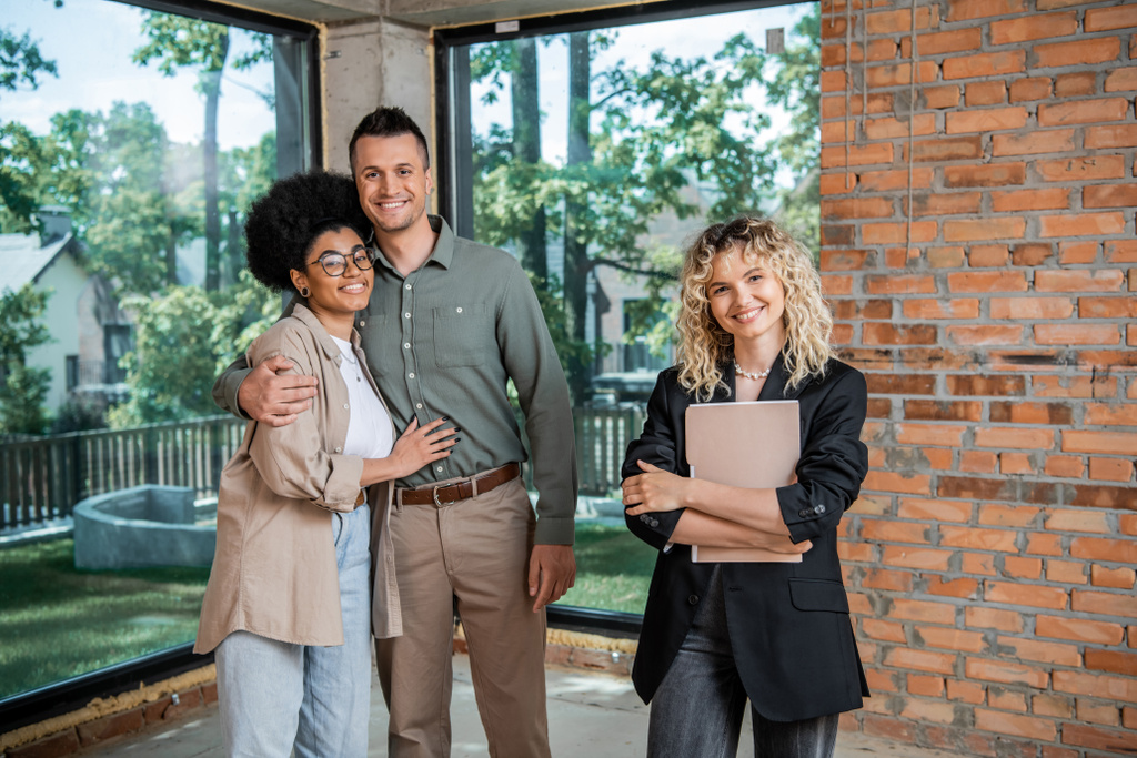 successful realtor with folder and cheerful multiethnic couple looking at camera in new cottage - Photo, Image