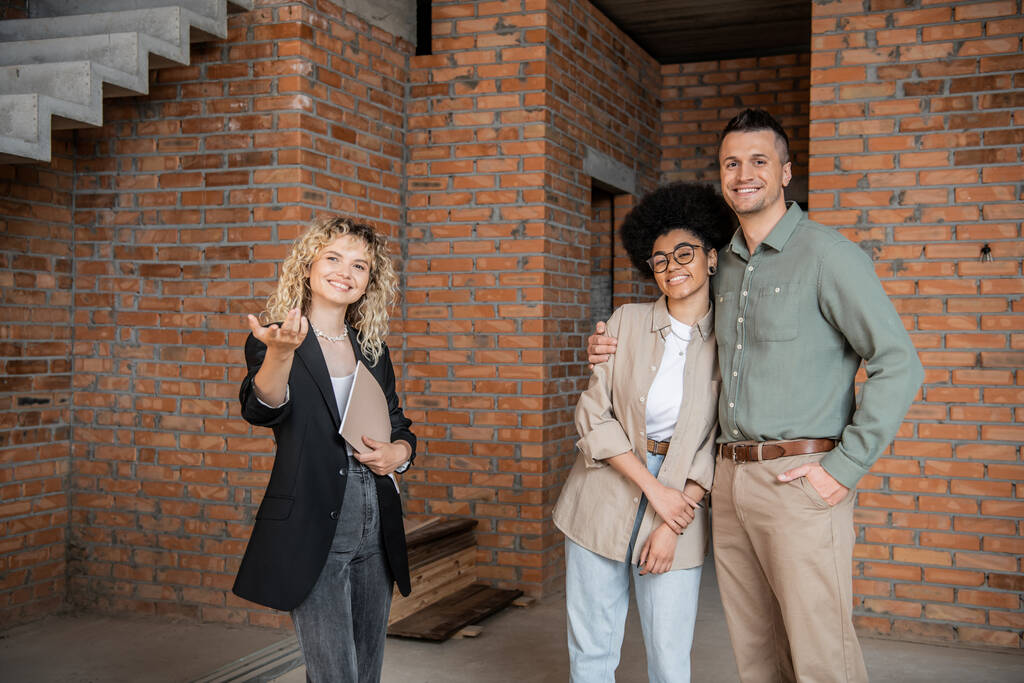 Lächelnder Immobilienmakler zeigt mit der Hand und zeigt multikulturellem Paar neue Wohnung - Foto, Bild