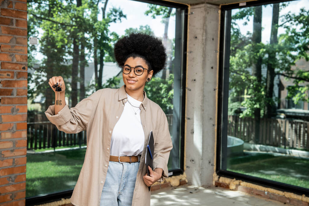 positiv afrikanisch-amerikanischer Immobilienmakler hält Ordner und Schlüssel in modernem Stadthäuschen - Foto, Bild