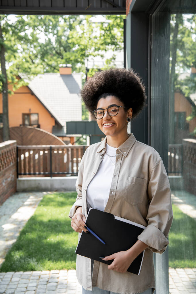 joyeux agent immobilier afro-américain avec des lunettes et un dossier regardant la caméra près de chalet - Photo, image