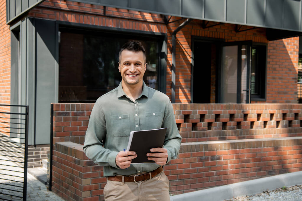 confident real estate agent holding folder and looking at camera near modern house on street - Photo, Image